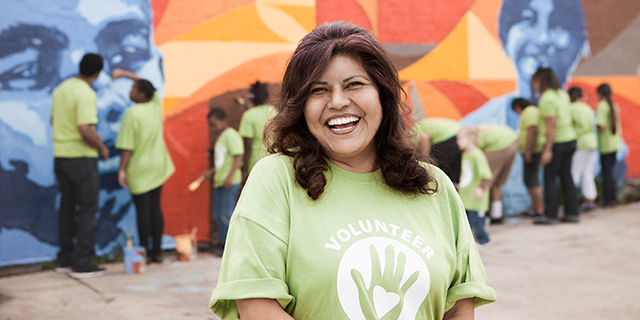 A smiling individual wearing a green "Volunteer" t-shirt stands in the foreground, exuding a sense of joy and enthusiasm. In the background, a group of people wearing similar t-shirts are collaboratively painting a vibrant mural on a wall, depicting community engagement and teamwork.